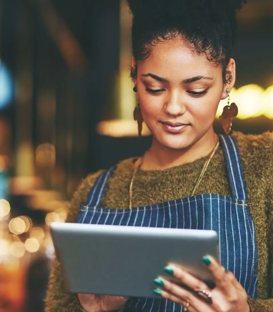 Frau in gestreifter Schürze benutzt ein Tablet in einem warm beleuchteten Raum, wie einem Café oder Restaurant